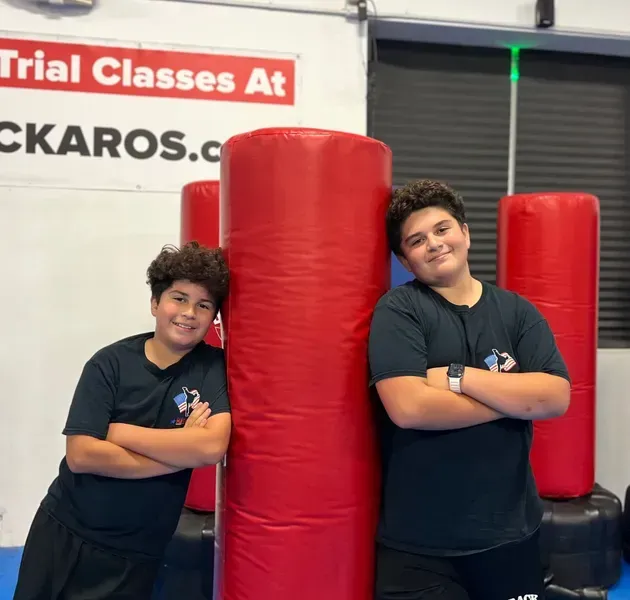 Two people in matching black shirts lean against large red punching bags in a gym, smiling with arms crossed.