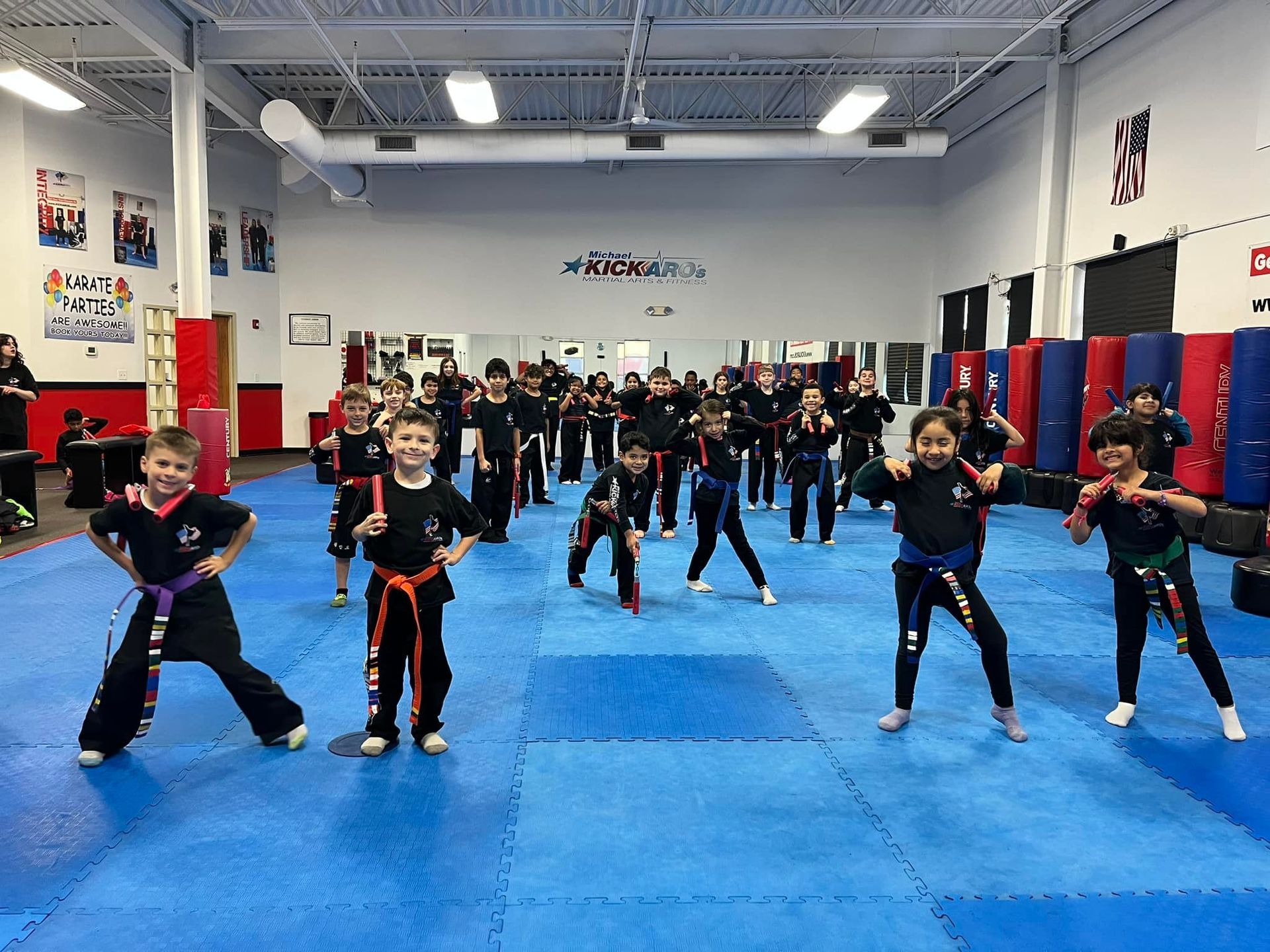 Kids in martial arts class on blue mats, wearing belts, holding training weapons, in a studio.