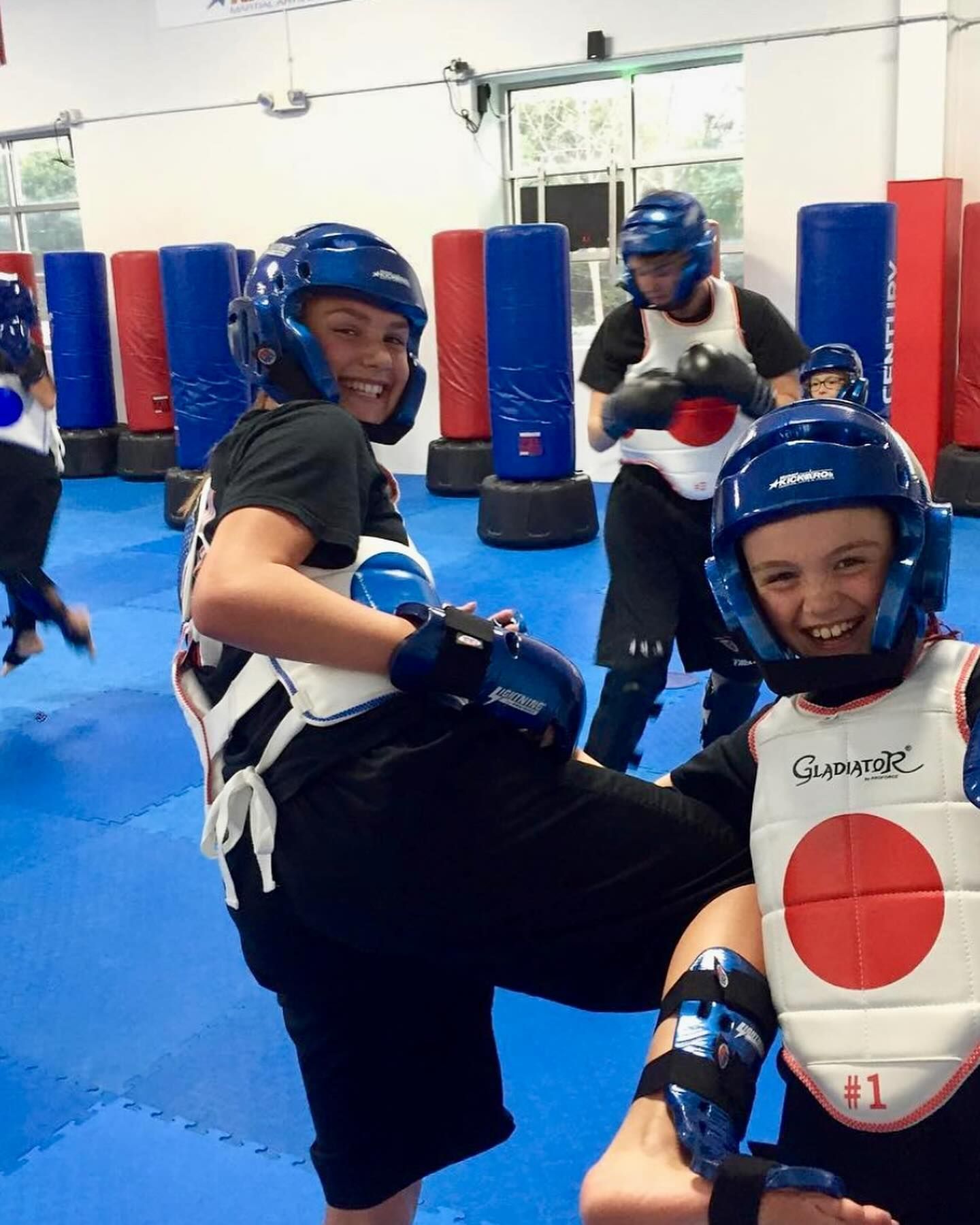 Kids in protective gear kicking in a training room. Blue mats, red punching bags, and smiles.