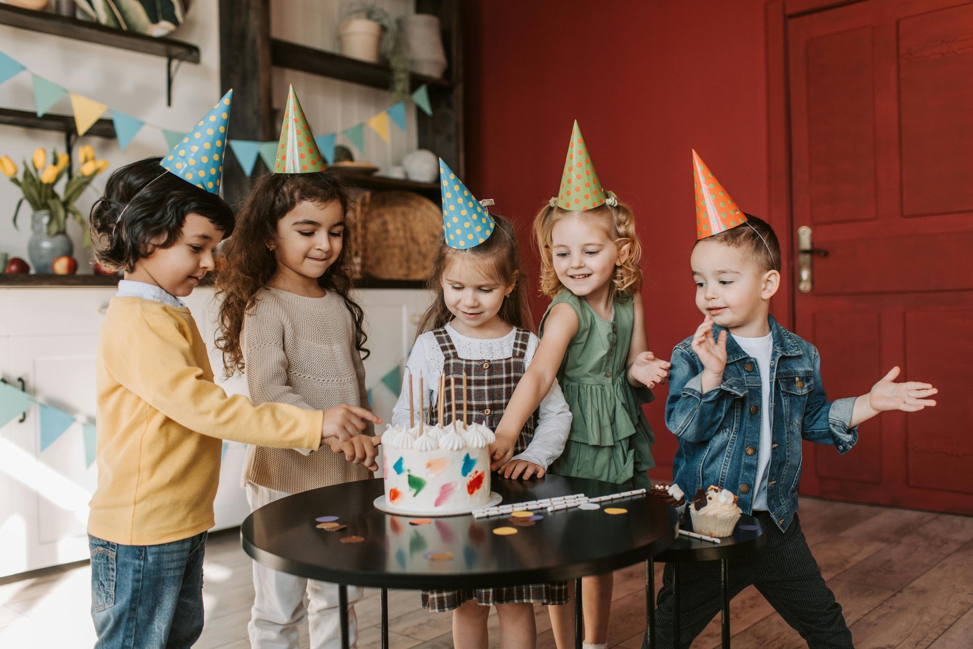 Five children in party hats gather around a cake, smiling at a birthday celebration.