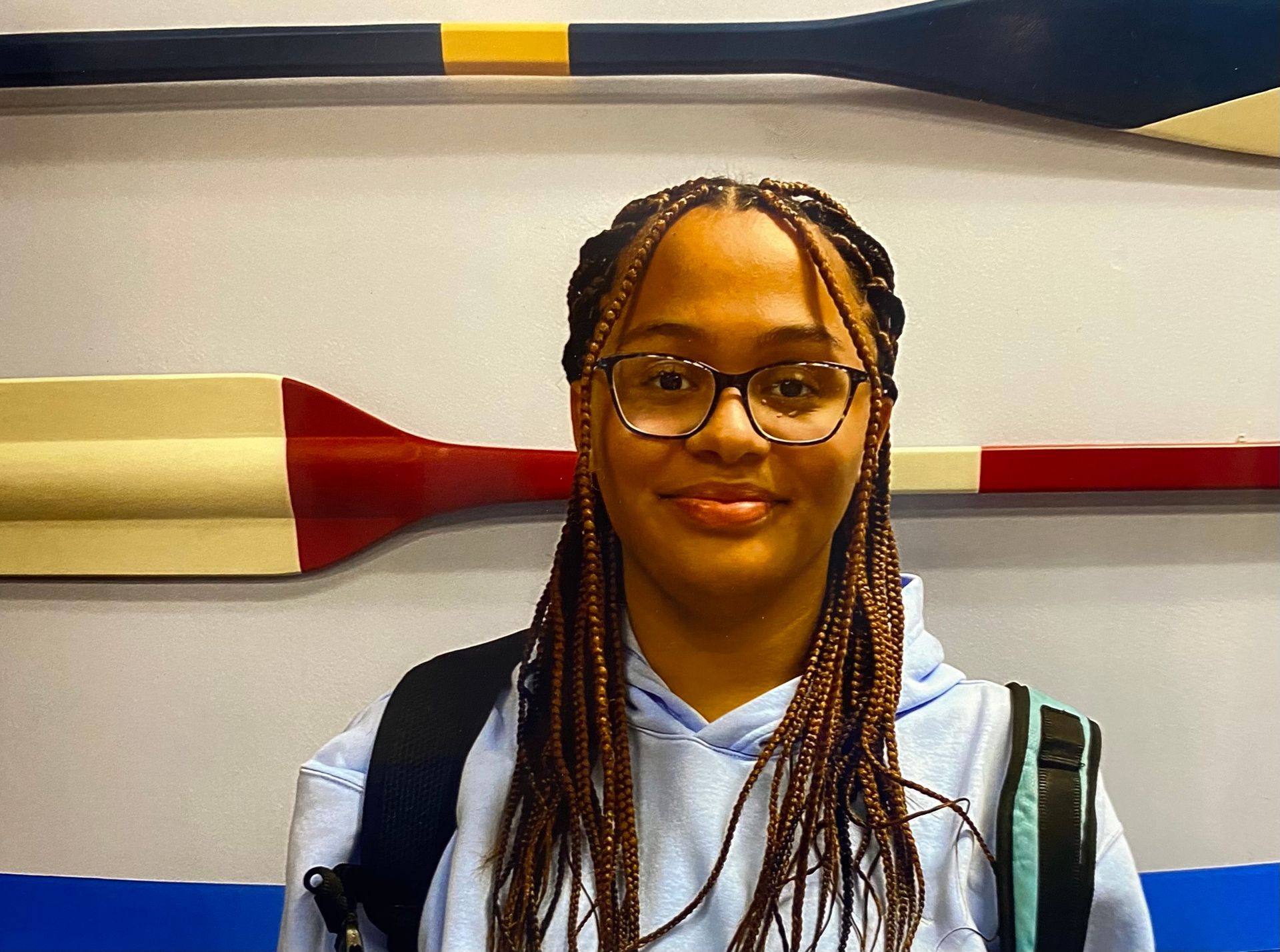 A woman wearing glasses is standing in front of a wall with oars on it.