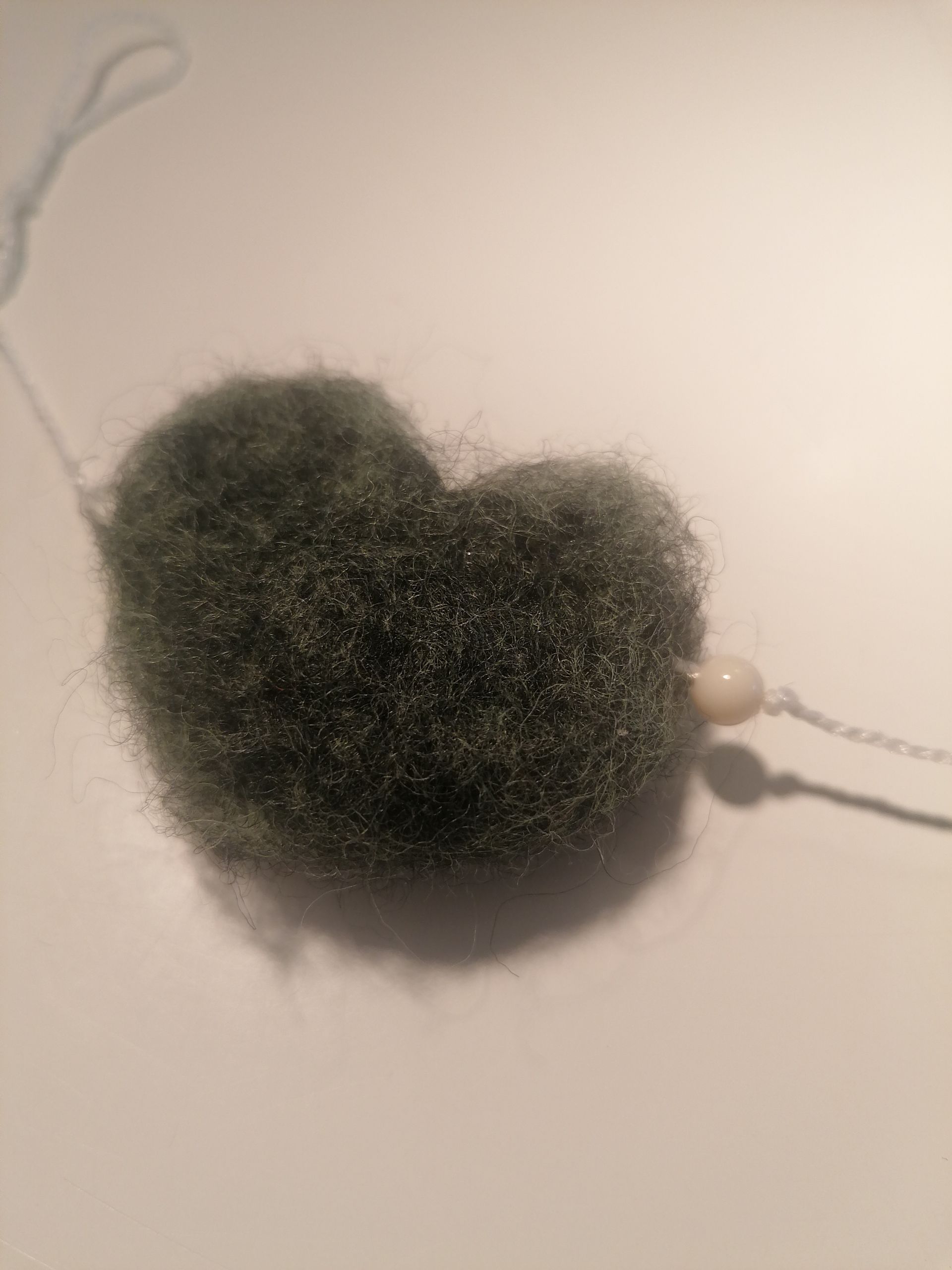 Groen, hartvormig, wol object met een witte kraal eraan.