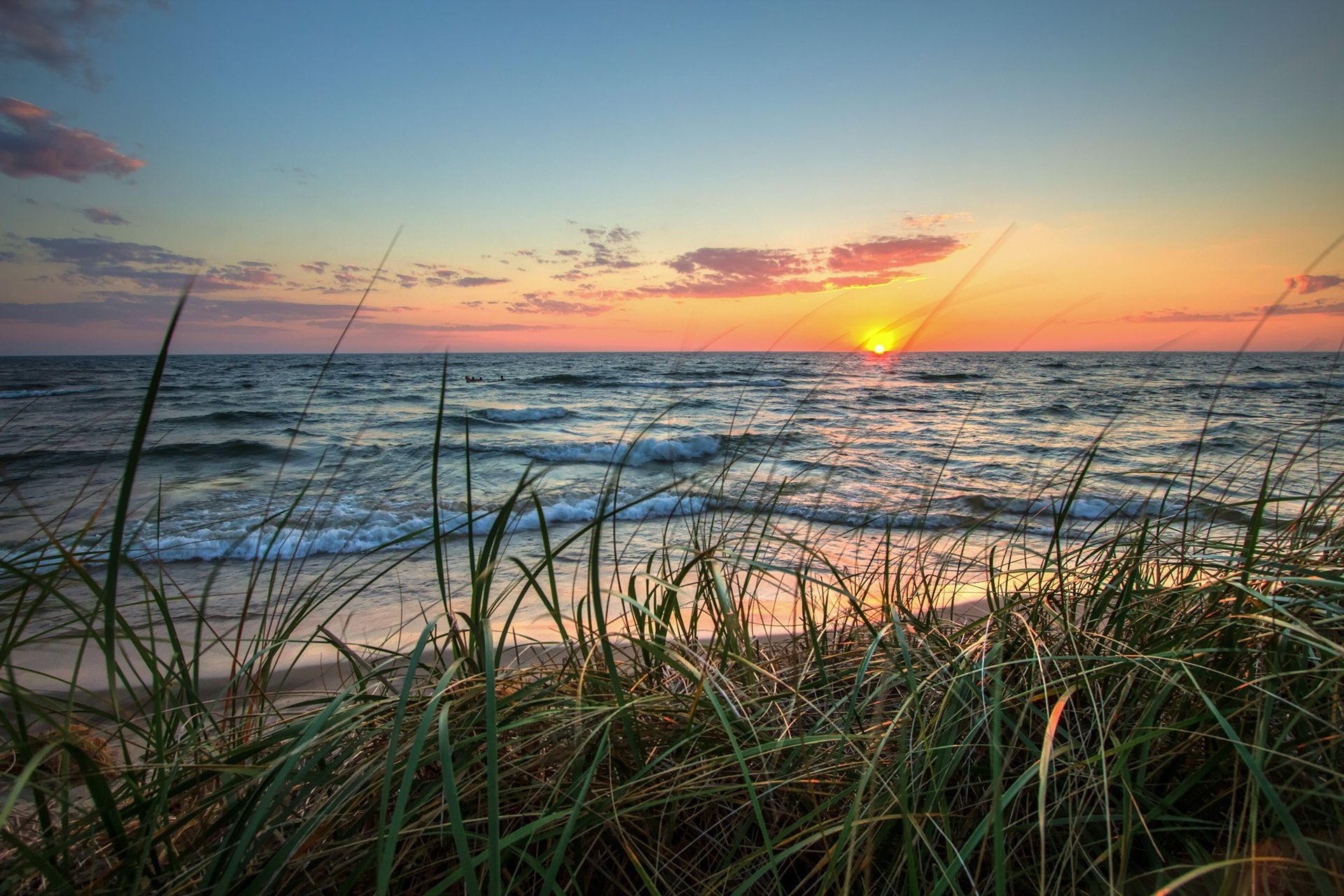 Sunset over a wavy ocean, seen from a sandy beach with green grass in the foreground.