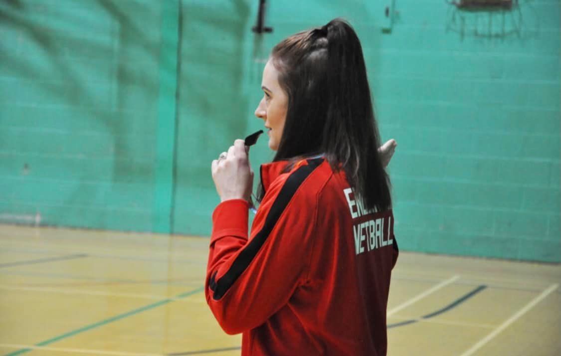 A woman in a red jacket is standing in a gym talking on a walkie talkie.