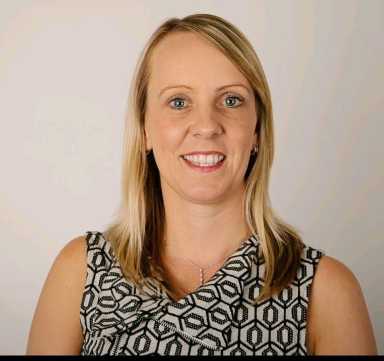 A woman in a blue shirt is standing in front of a wall of white flowers.