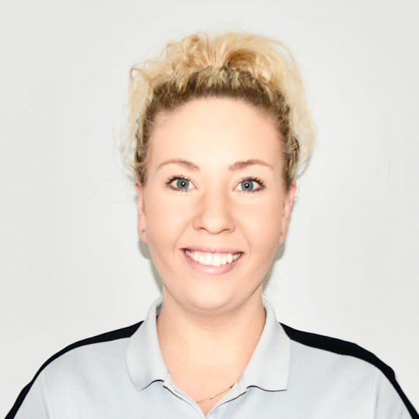 A woman in a blue polka dot dress is smiling in front of a wall of white flowers.