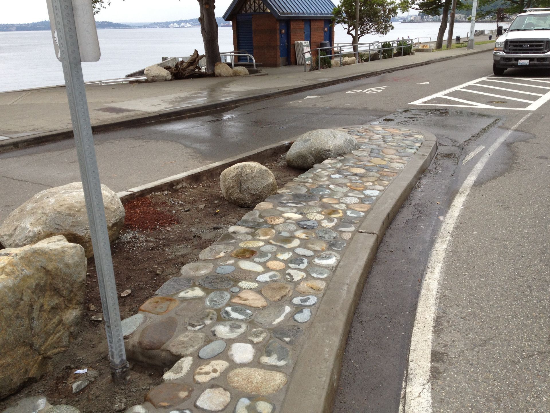 Curved stone curb along a wet waterfront road, with rocks, a small shelter, and a parked car