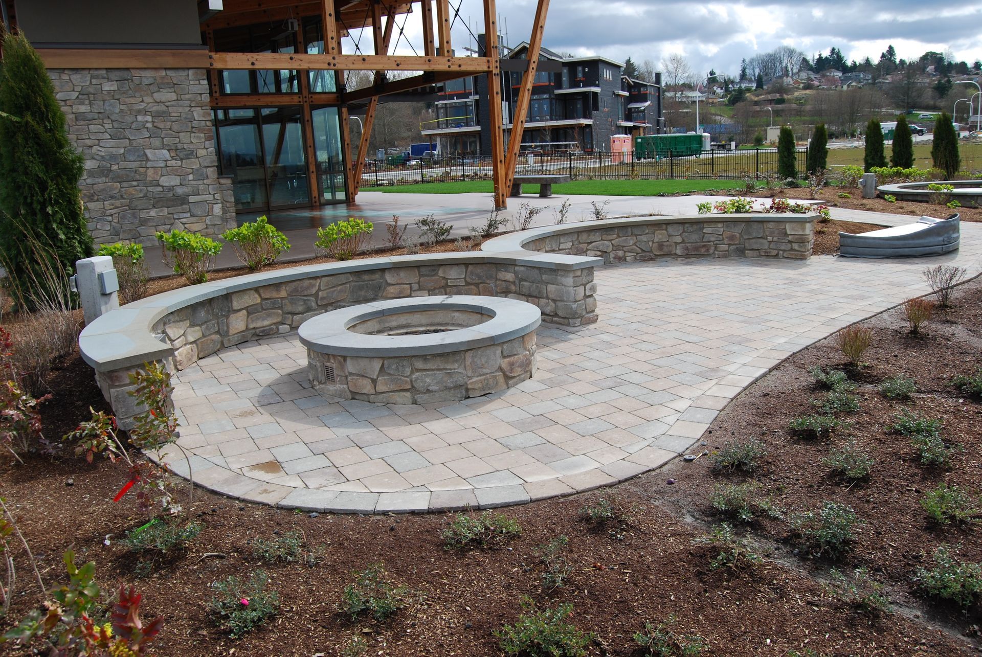 Curved stone patio with built-in circular fire pit and benches beside a modern building and landscaped garden