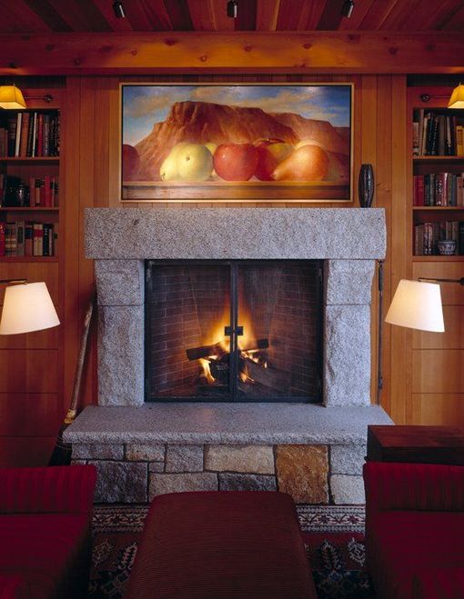 Cozy living room fireplace with a lit fire, stone mantel, wood-paneled walls, and a landscape painting above.
