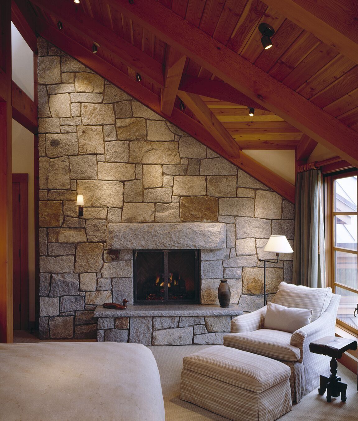 Stone fireplace in a cozy living room with sloped wood ceiling and beige armchair by a window.