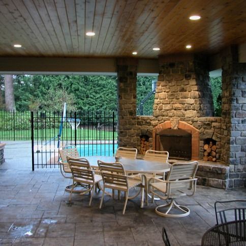 Covered patio with stone fireplace, white table and chairs, and a pool view beyond a gate