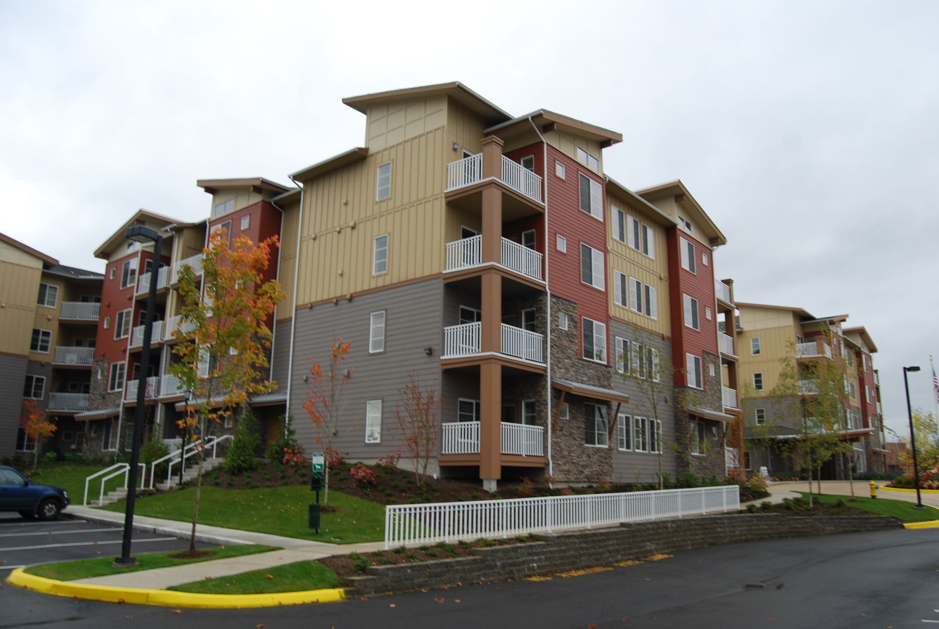 Modern apartment buildings on a street corner with balconies, autumn trees, and overcast sky
