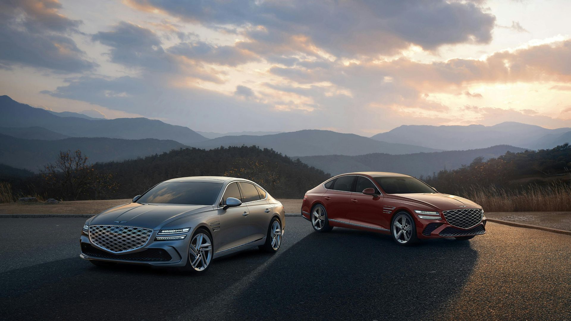 Two sleek sedans, silver and red, parked on a road at sunset with mountains in the background