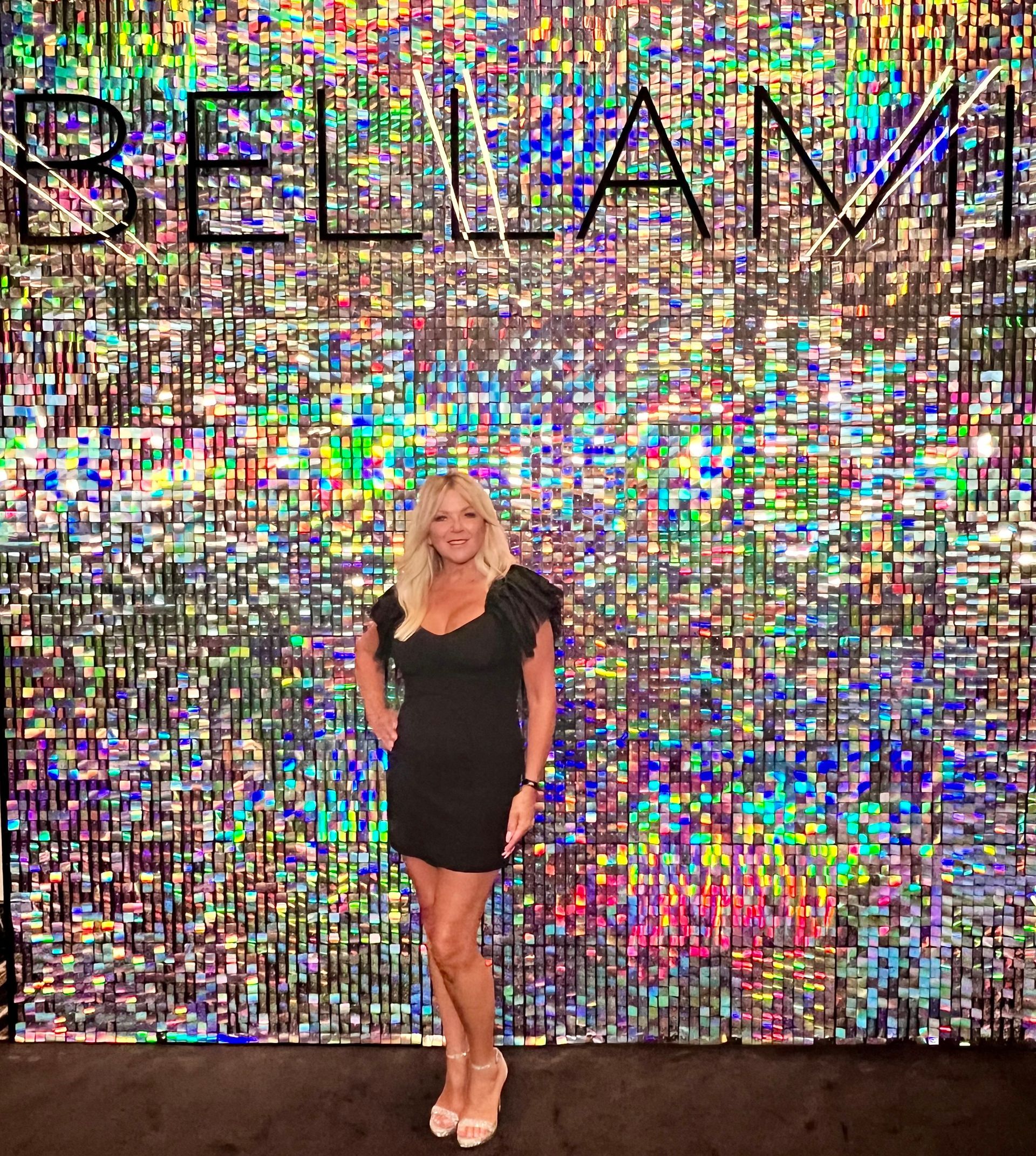Woman with blonde hair sits in black chair, wearing black outfit, in hair salon.
