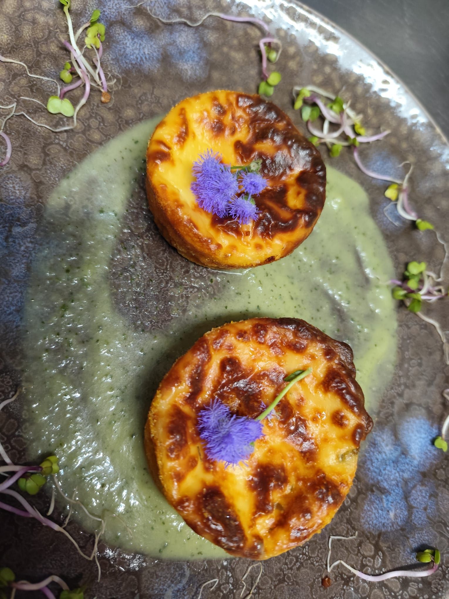 Due tortine salate dorate su salsa verde, guarnite con fiori viola e germogli.
