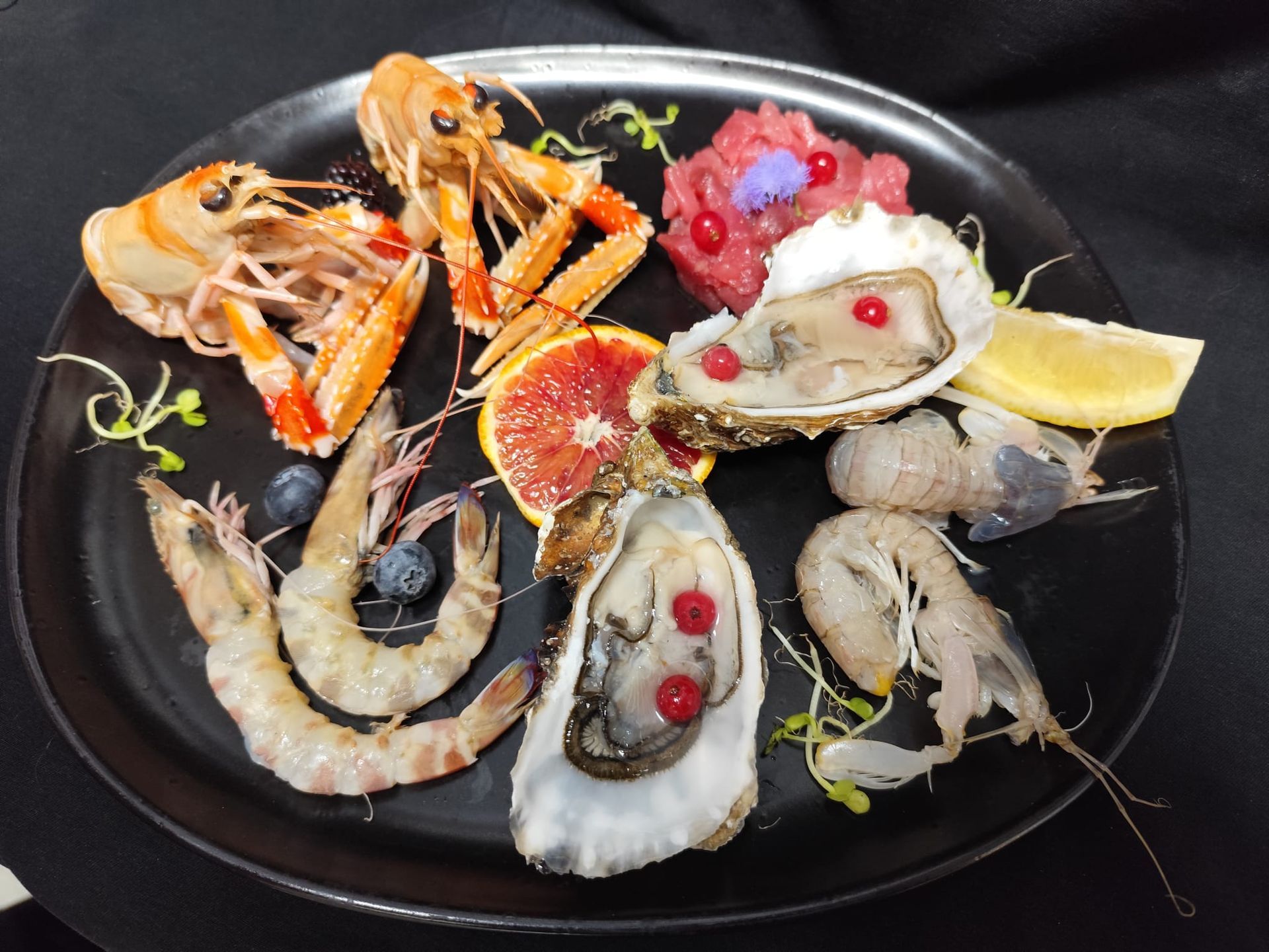 Piatto di frutti di mare: scampi, gamberi, ostriche, una fetta di agrumi e guarnizioni su un piatto scuro.