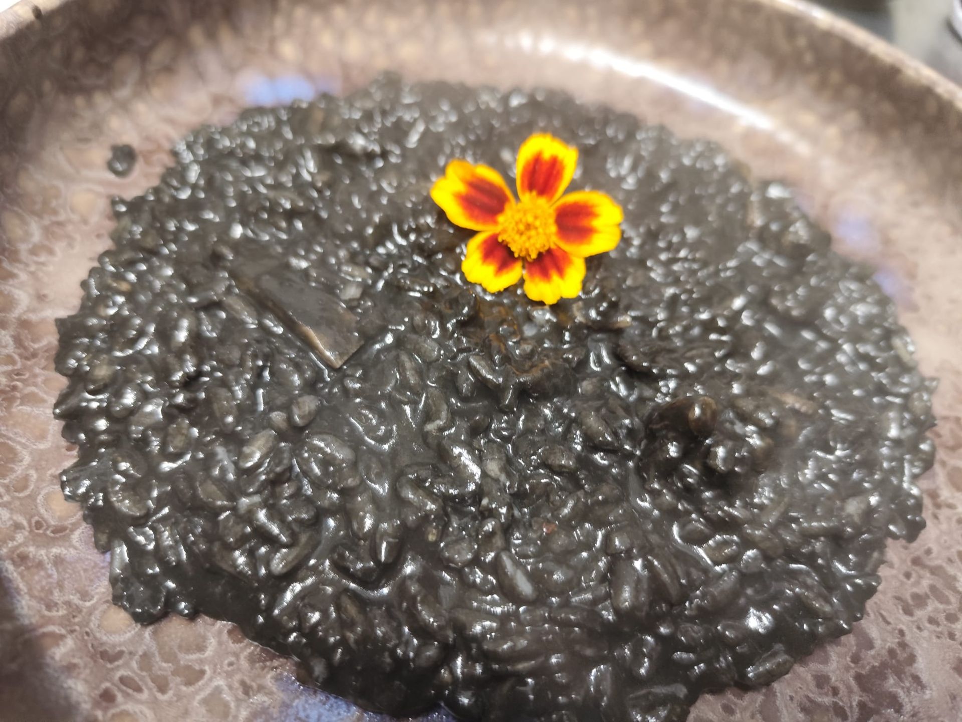 Risotto nero guarnito con un fiore giallo e arancione su un piatto marrone maculato.
