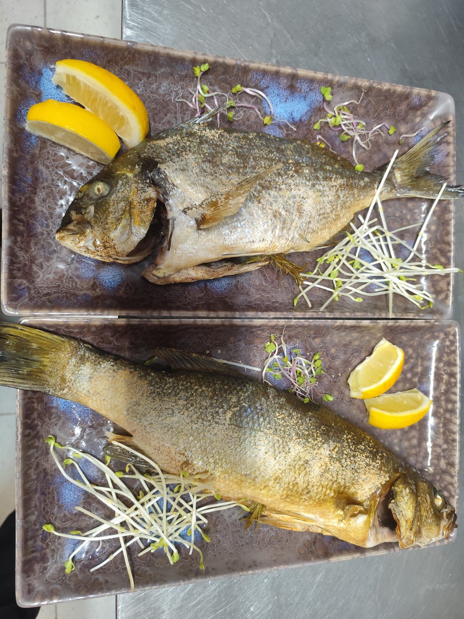 Due pesci interi dorati, serviti su piatti rettangolari decorativi, guarniti con fette di limone e germogli.