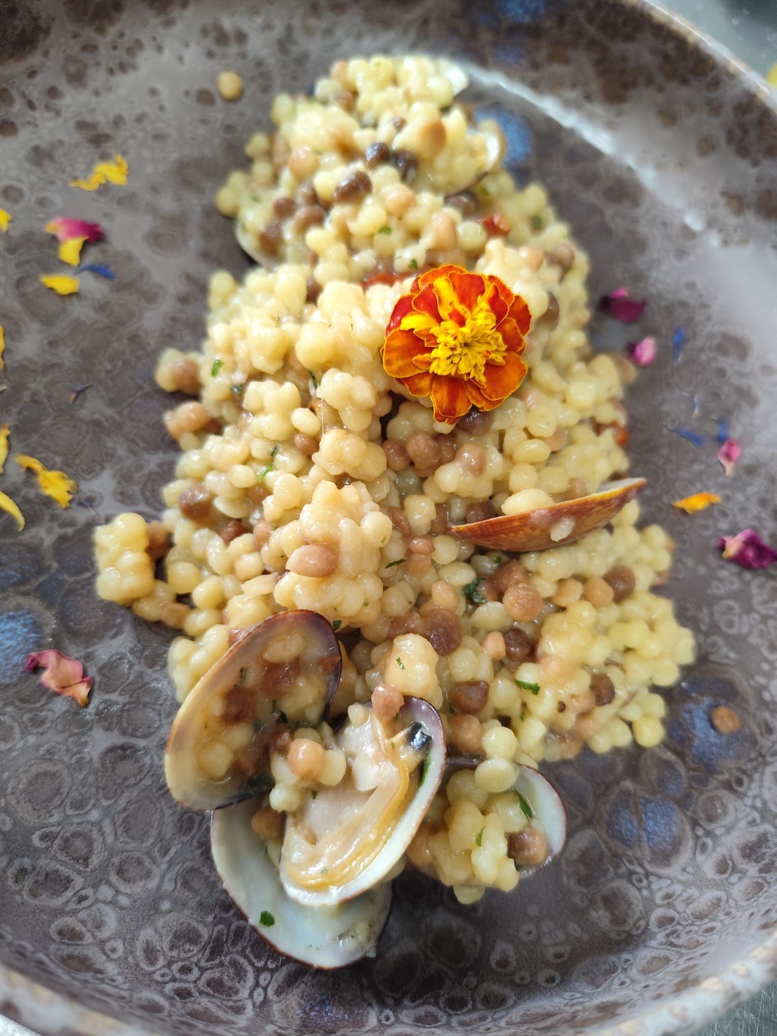 Piatto di couscous con vongole, lenticchie, fiori commestibili ed erbe aromatiche in una ciotola marrone screziata.