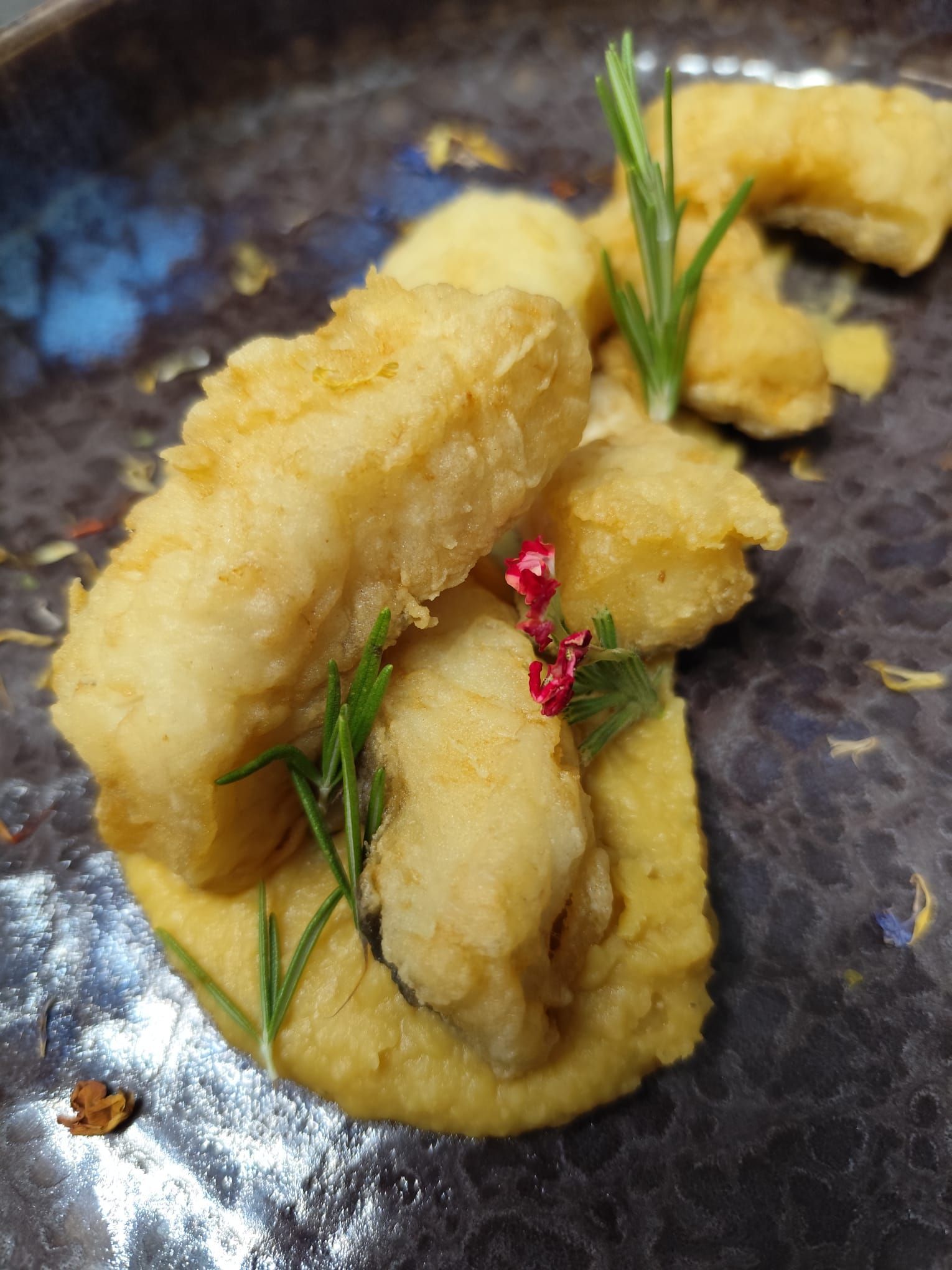 Cibo fritto su purea gialla, guarnito con rosmarino e piccoli fiori rosa, su un piatto scuro.