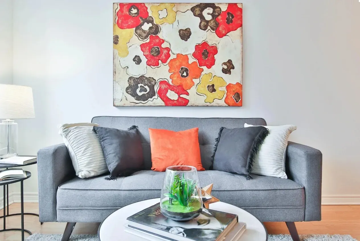 A modern living room featuring a comfortable grey sofa with colorful pillows, a round coffee table, and a vibrant floral painting.