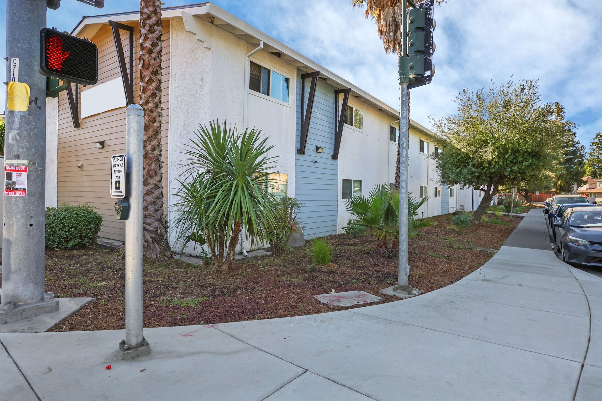 Apartment building corner with sidewalk, palm trees, parked cars, and pedestrian signal.