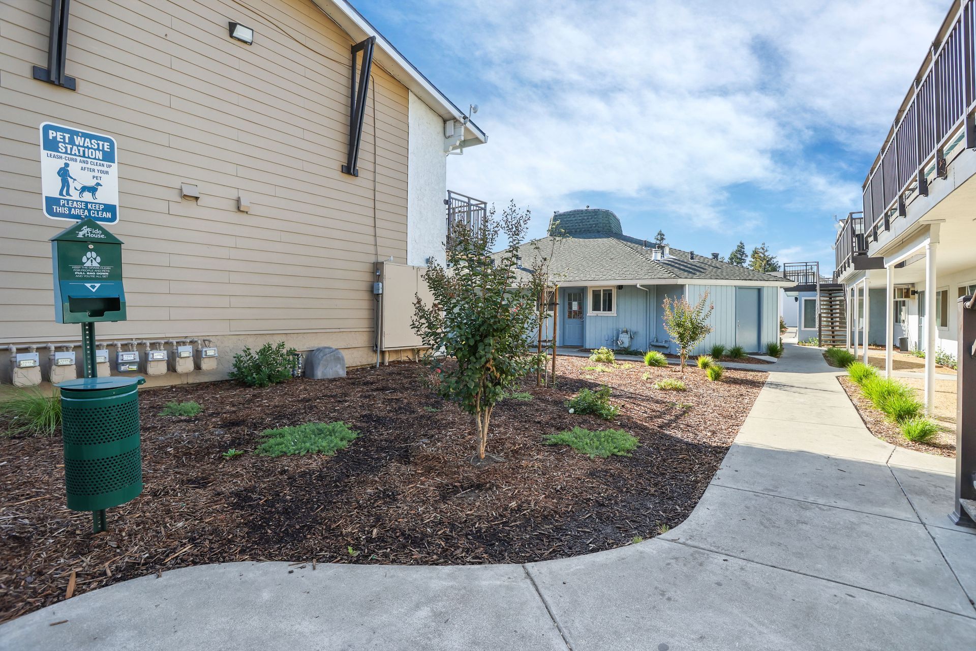 Apartment courtyard with beige buildings, landscaped mulch beds, and a concrete walkway under a blue sky