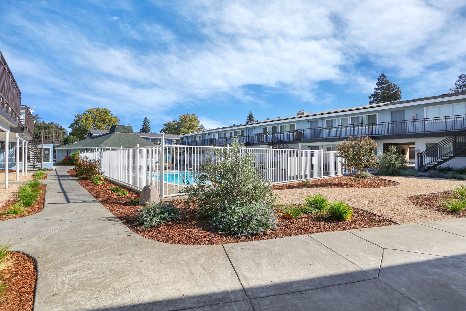 Apartment courtyard with pool, white fence, landscaping, and row houses under a blue sky