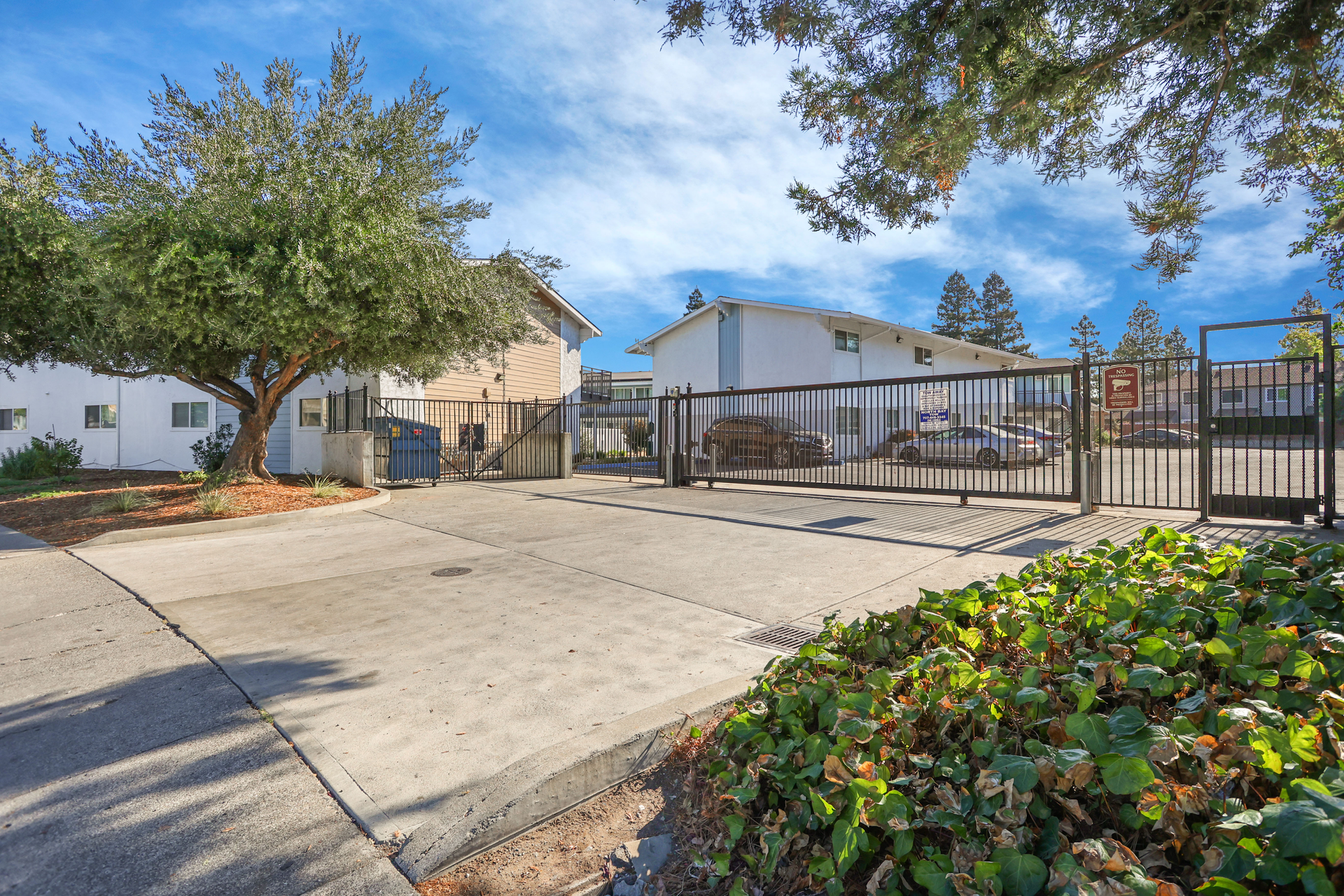 Gated apartment courtyard with concrete path, trees, and landscaped bushes under a blue sky