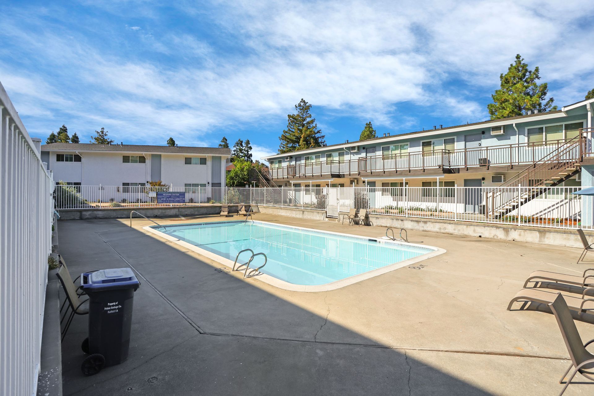 Motel courtyard with a swimming pool, two-story rooms, lounge chairs, and blue sky.