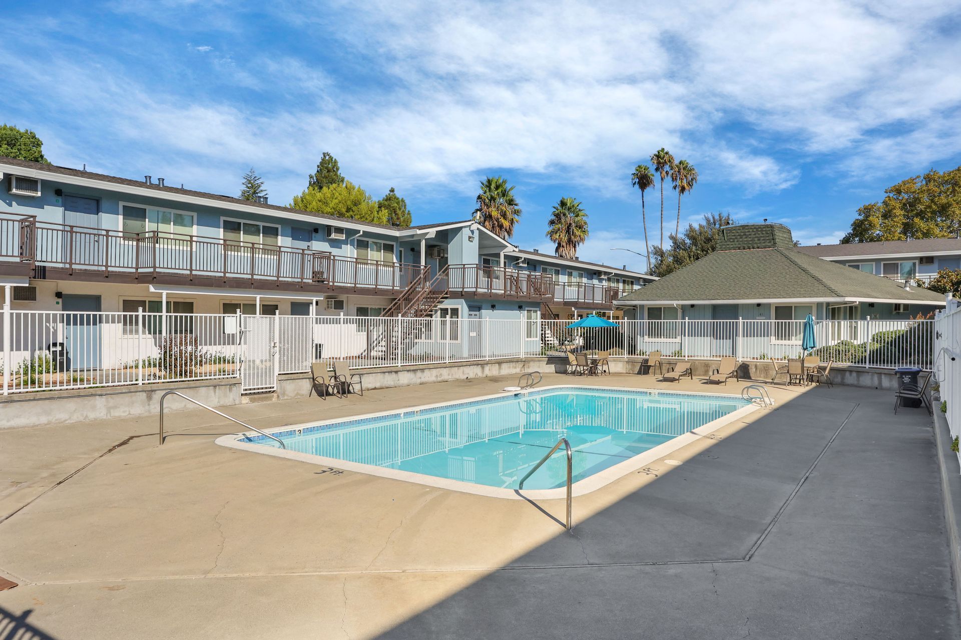 Motel courtyard with outdoor swimming pool, two-story rooms, and palm trees under a blue sky