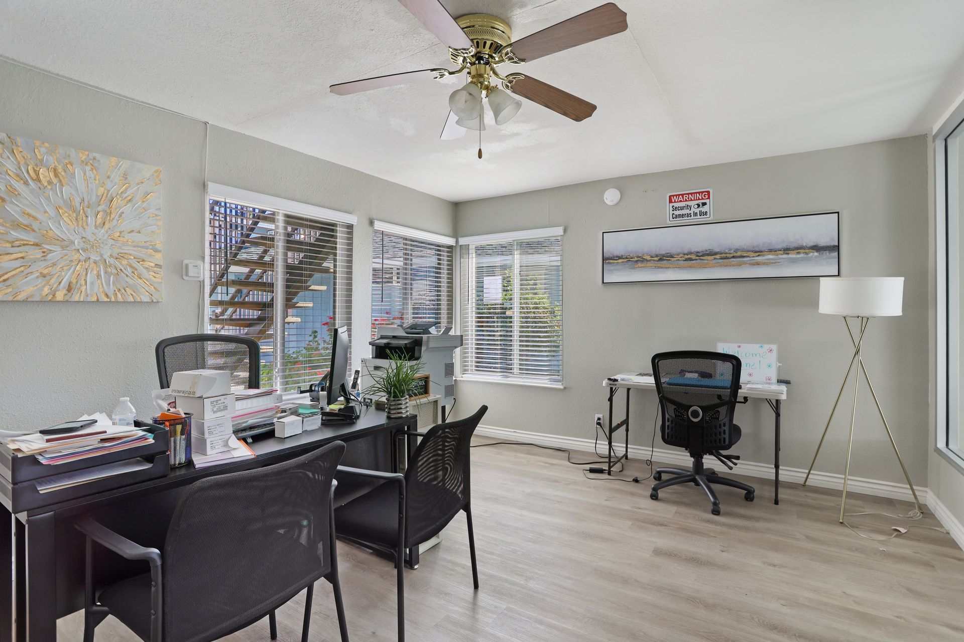 Home office with two desks, black chairs, a floor lamp, and beach wall art in a bright room.