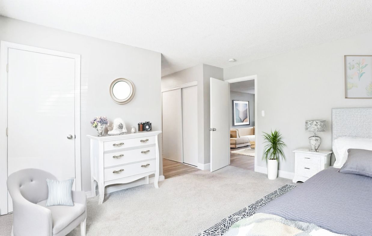 Bright white bedroom with a dresser, mirror, chair, plant, and bed with gray bedding.