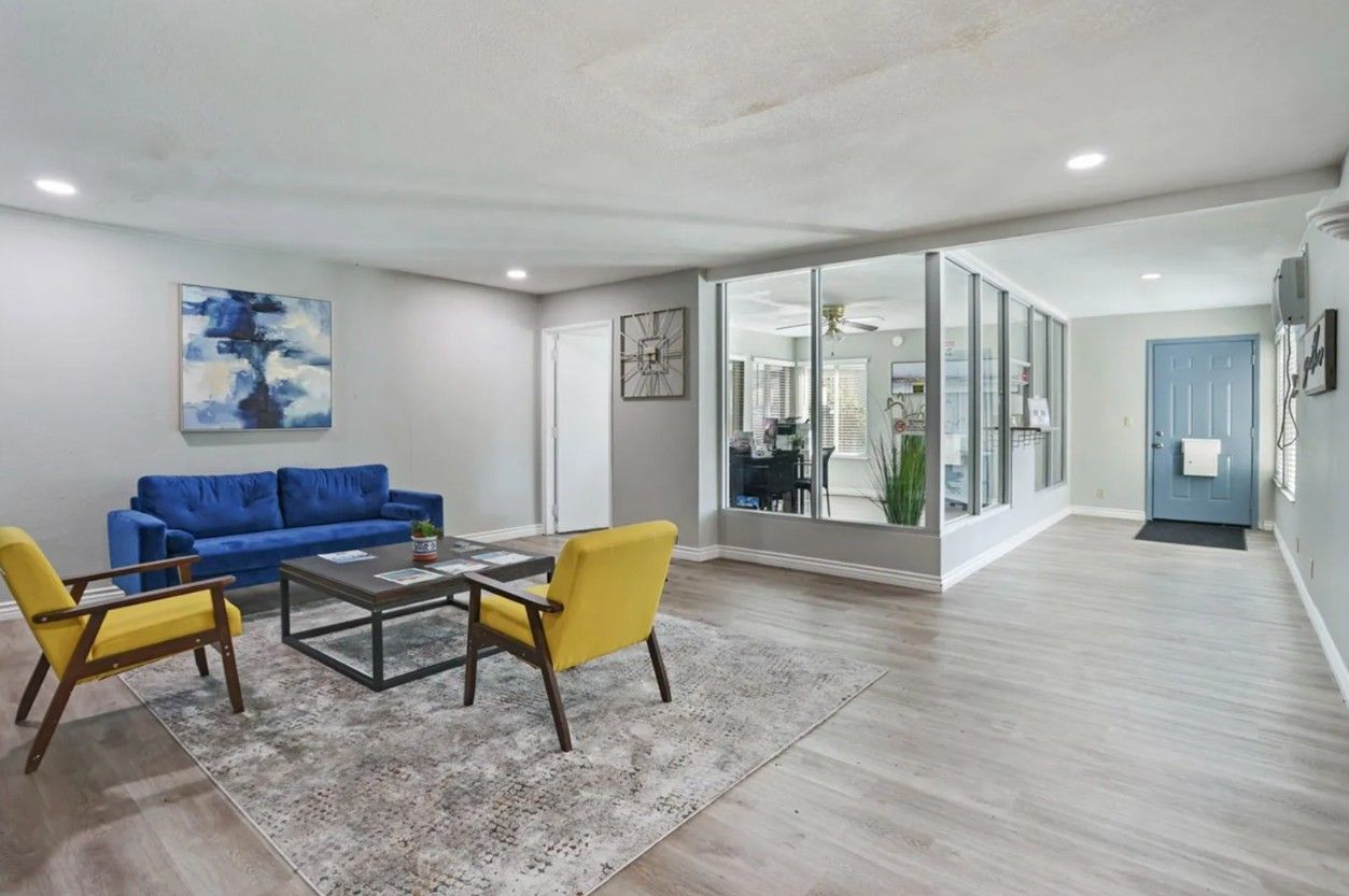 Modern lounge with blue sofa, yellow chairs, glass-walled room, and light wood flooring