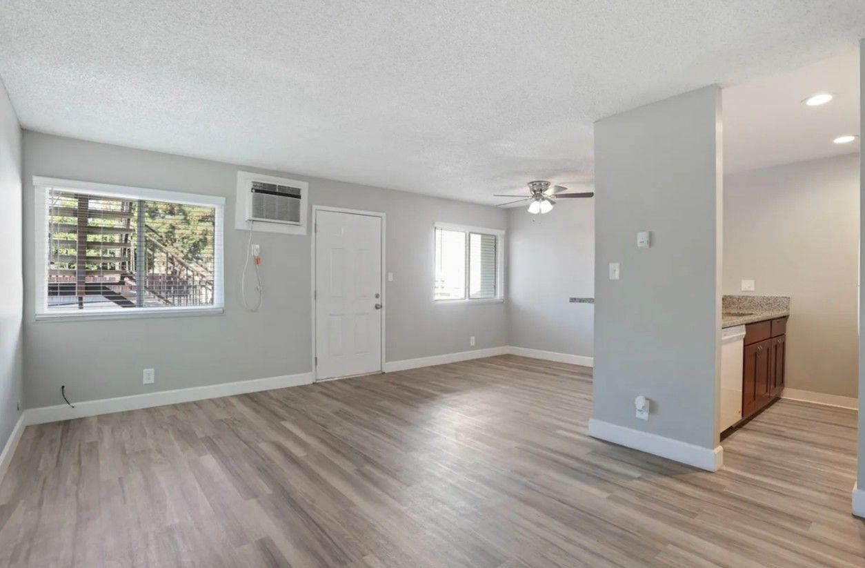 Empty gray living room with wood floors, white doors, and open view into a kitchen area