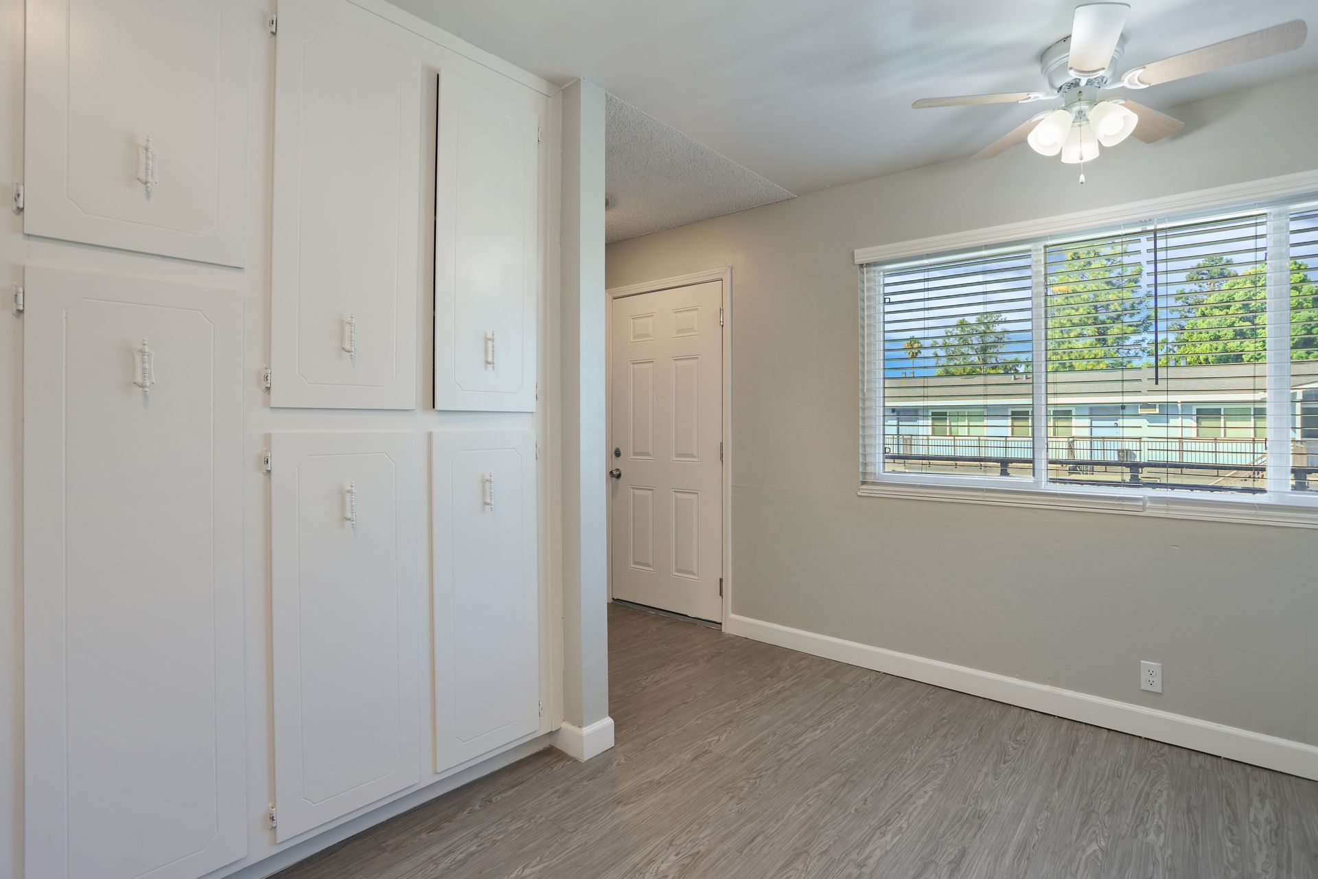 Bright empty bedroom with white built-in cabinets, gray carpet, and a window with blinds and a ceiling fan.