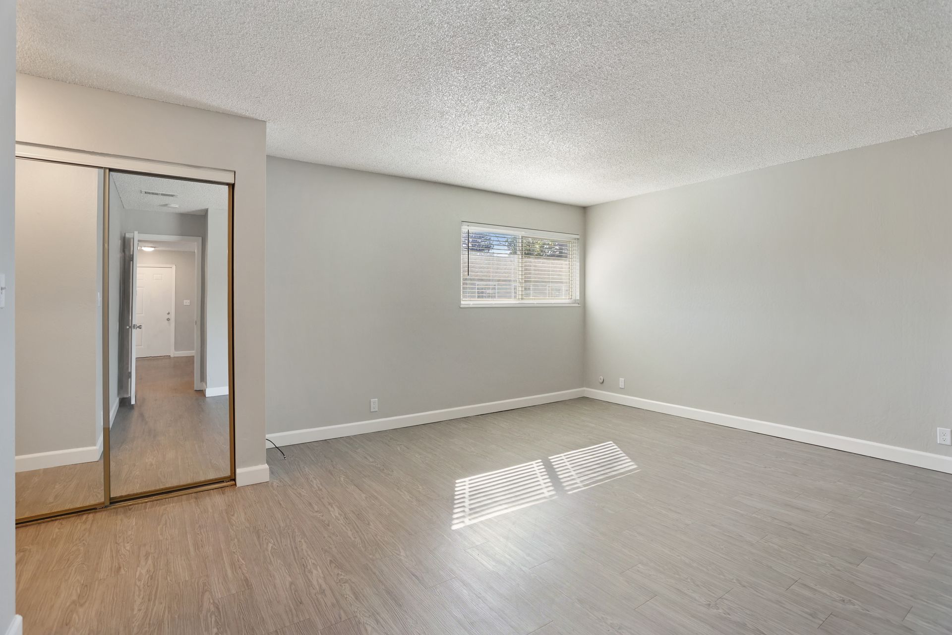 Empty beige room with gray flooring, mirrored closet door, and a small window.