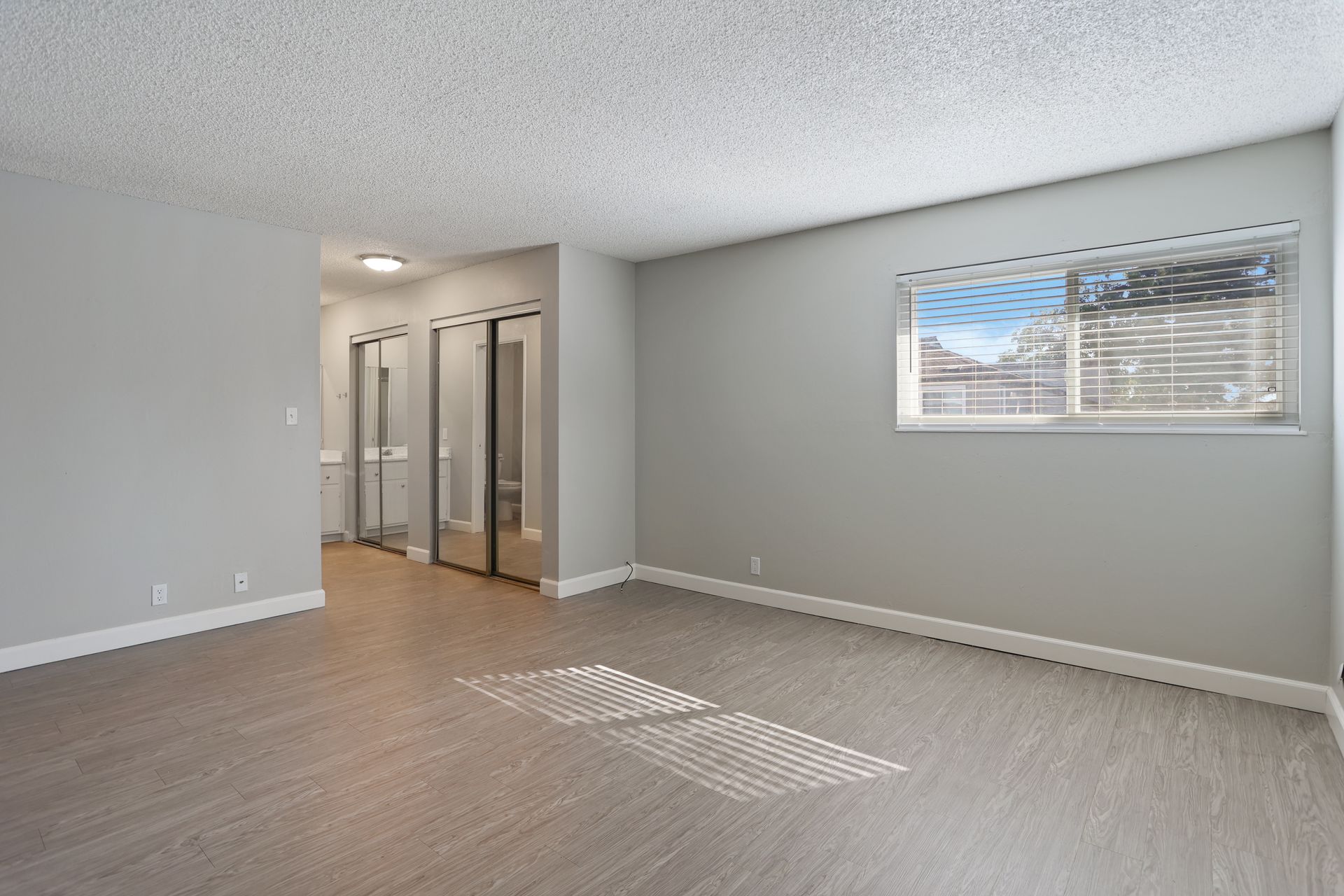 Empty beige bedroom with carpet, closet doors, and a small window letting in sunlight.