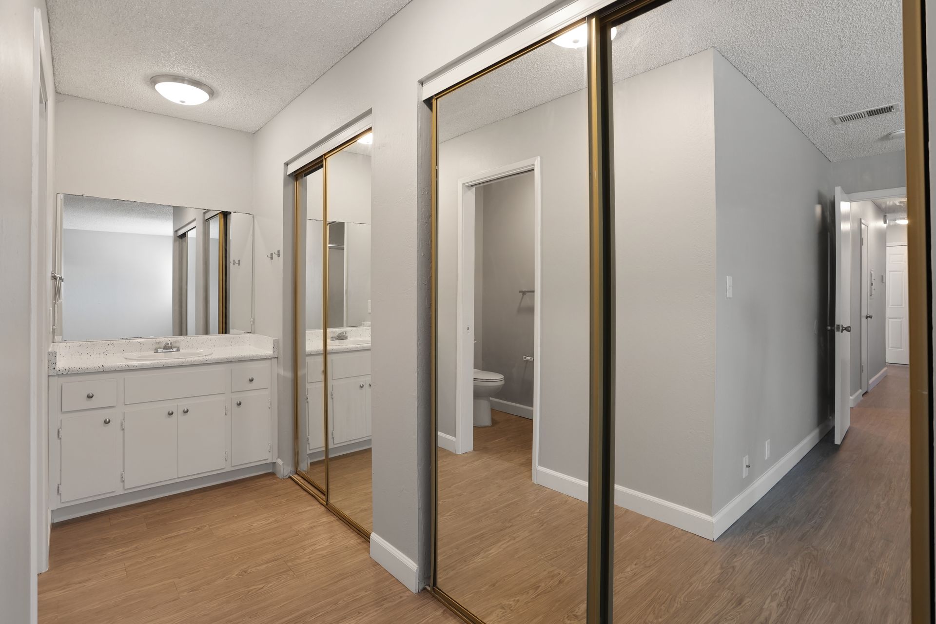 Bright bathroom with vanity, mirrors, glass shower, and white walls beside a hall with wood flooring