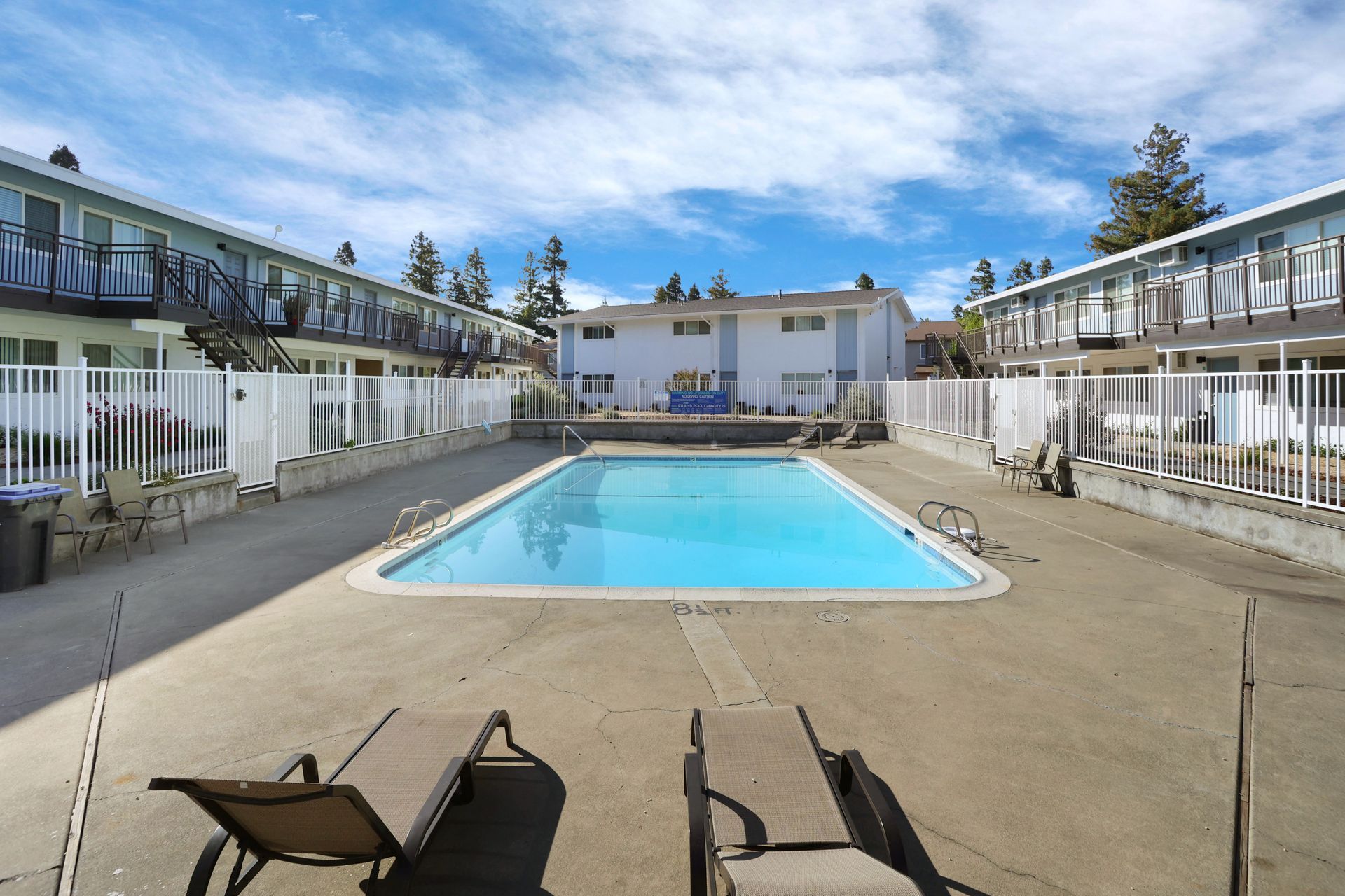 Outdoor motel courtyard with a central swimming pool, white fencing, and lounge chairs under a blue sky