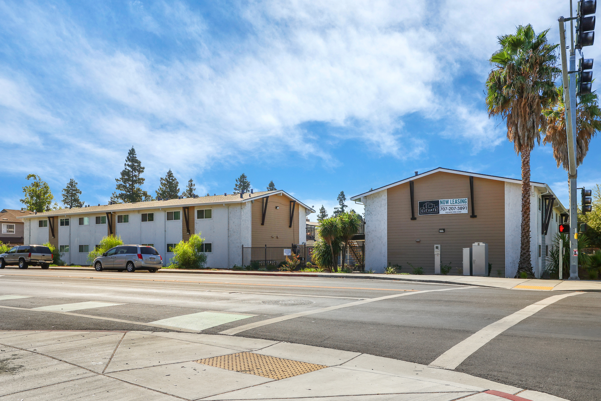 Street corner with low beige apartment buildings, palm trees, and a blue sky with clouds.