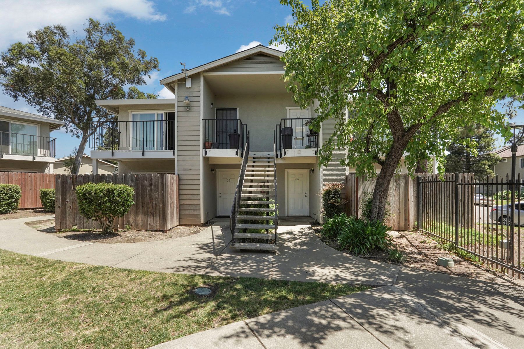 Two-story apartment building with exterior stairs, fenced yard, and trees in front