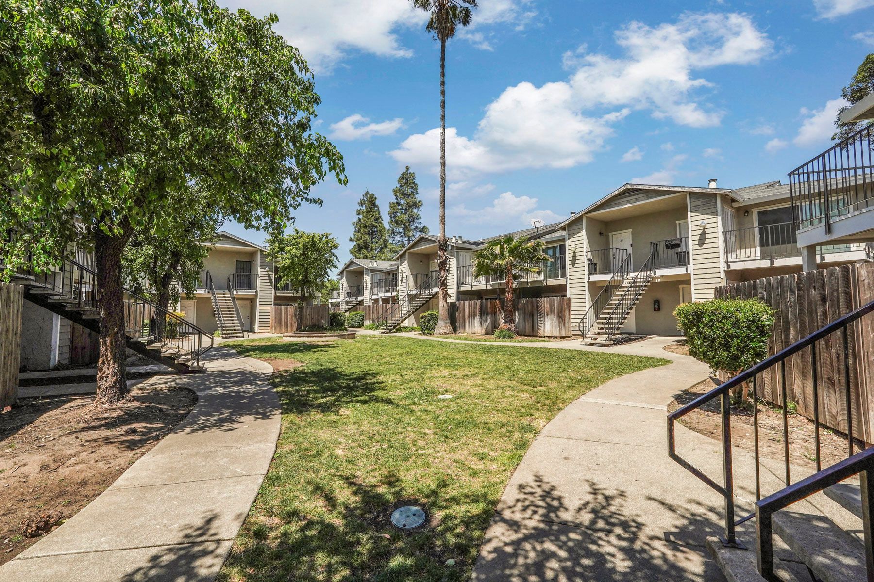 Apartment courtyard with green lawn, walkways, trees, and beige buildings under a blue sky