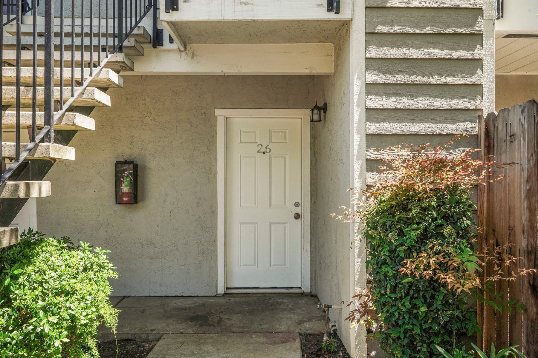 Exterior apartment entryway with white door, stairs, and shrubs in sunlight