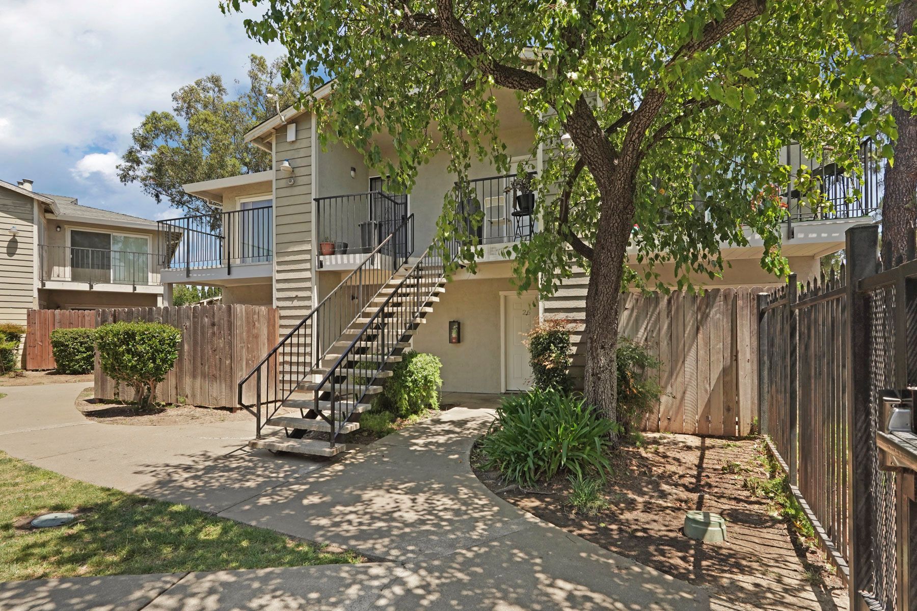 Apartment exterior with stairs, balcony, and tree-shaded yard by wooden fence