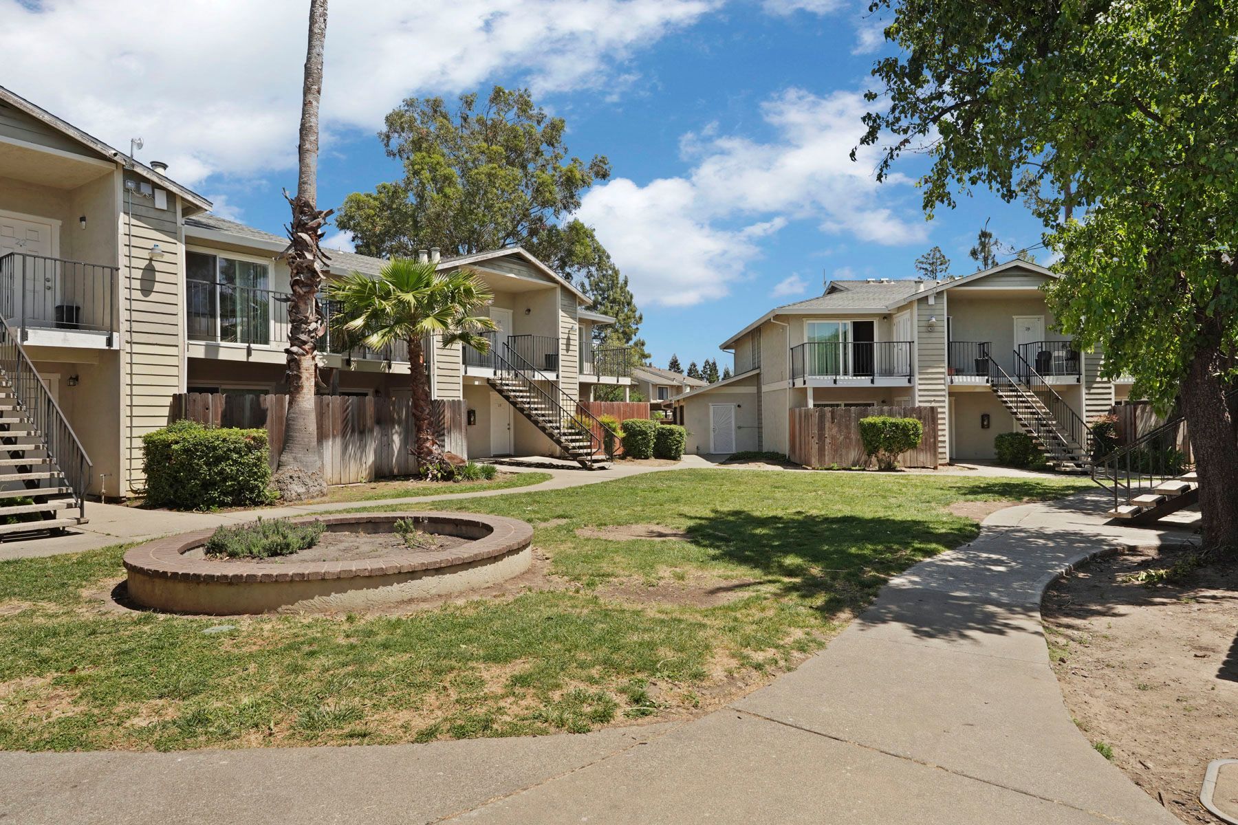 Apartment courtyard with grass, trees, walkways, and beige two-story buildings under a blue sky