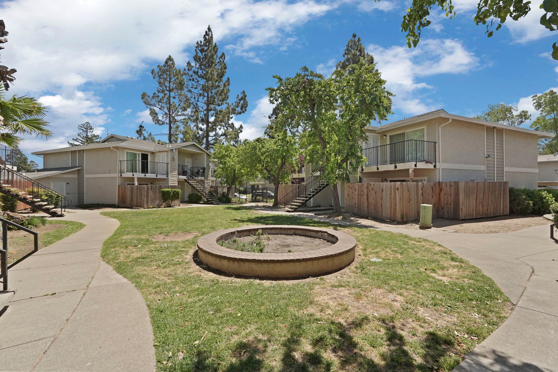 Apartment courtyard with a circular fire pit, grass, sidewalks, trees, and homes under a blue sky