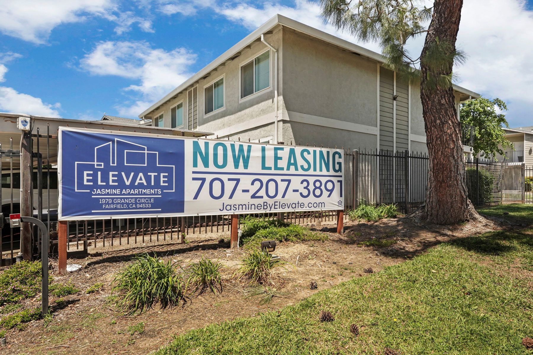 Apartment building with “Now Leasing” sign and phone number, beside a fence and palm trees