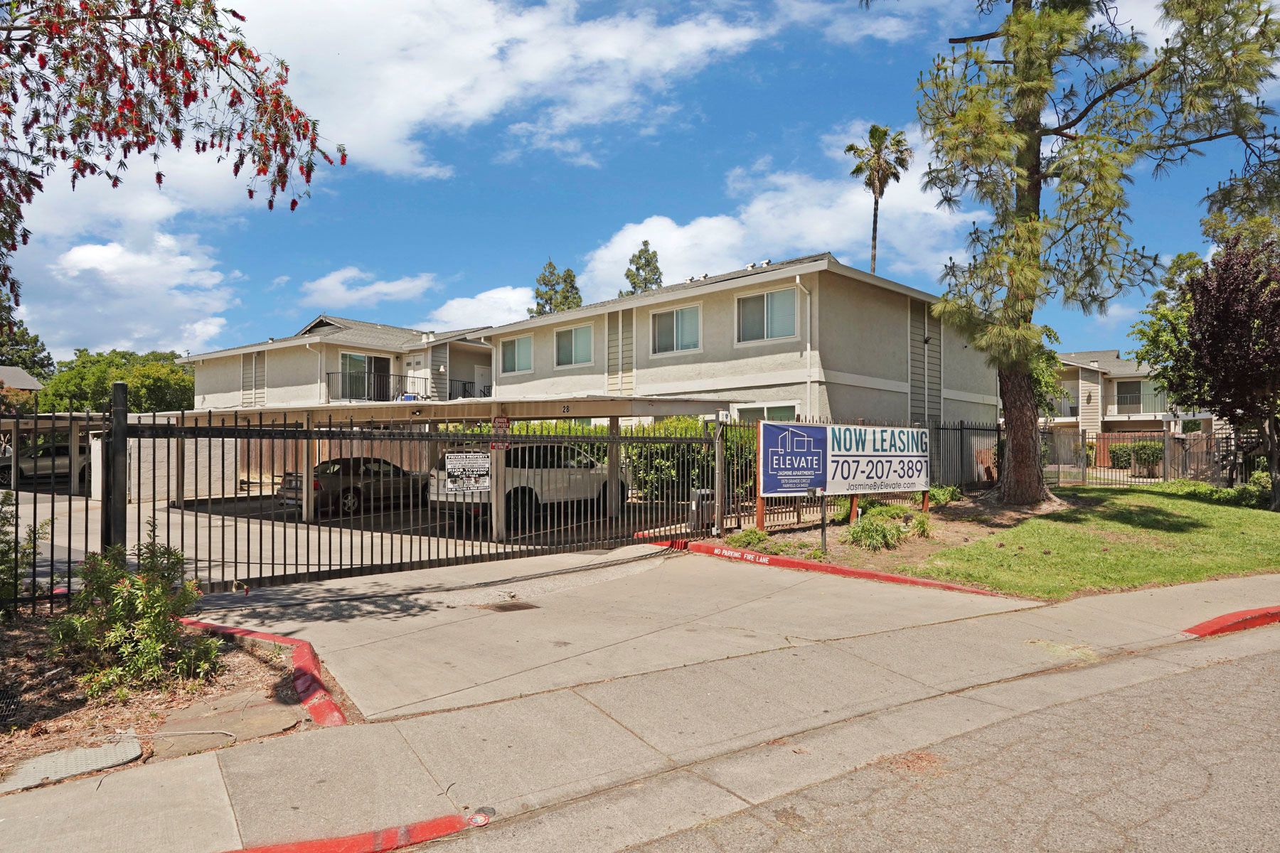 Apartment complex entrance with gated driveway, beige buildings, trees, and a blue sign on a sunny day