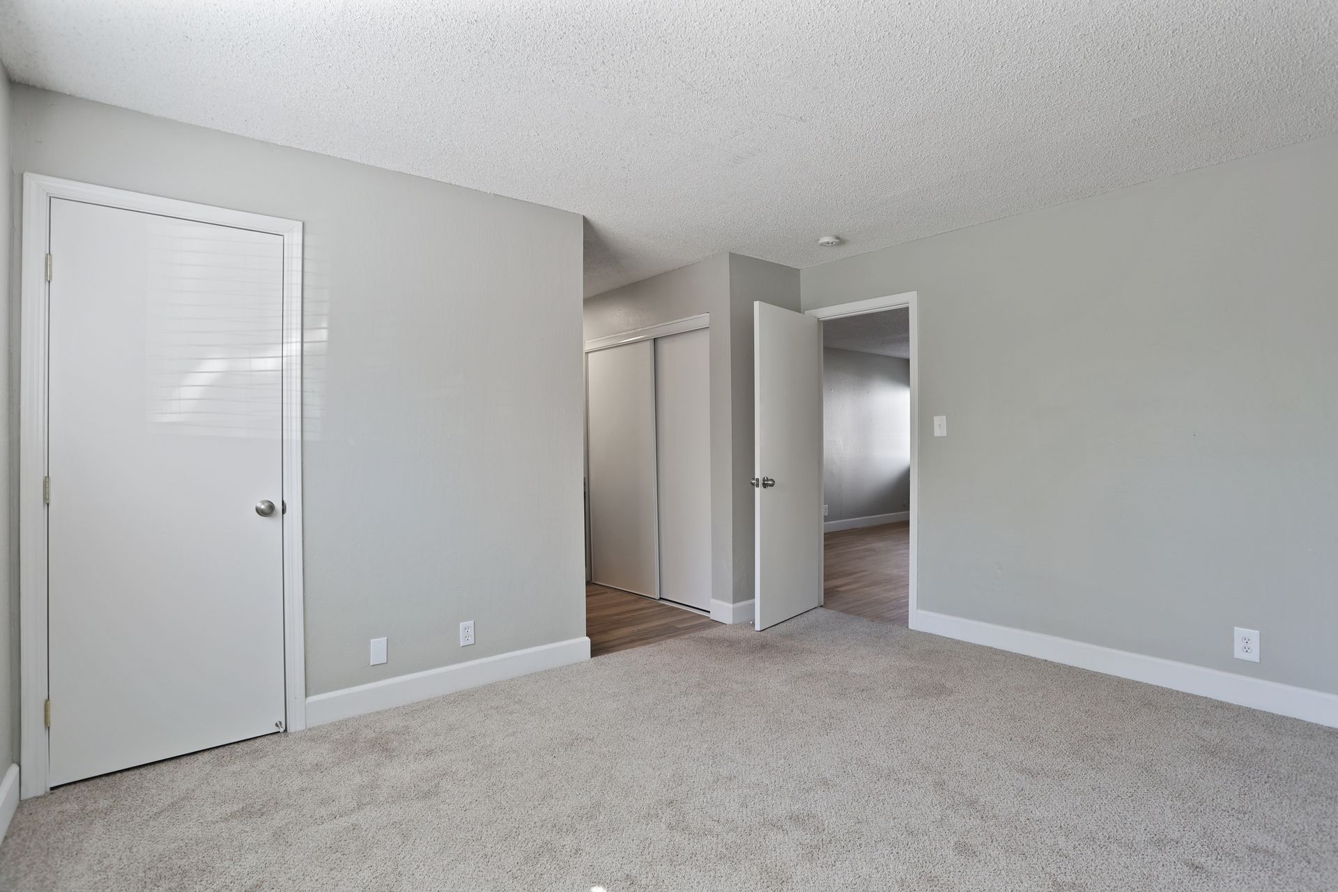Empty beige bedroom with carpet, white closet doors, and an open doorway to a hall.